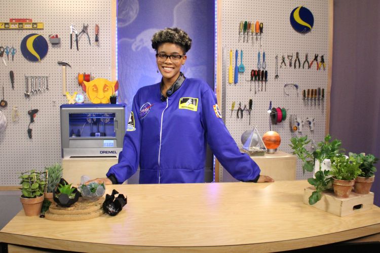 Netia McCray is shown wearing a blue jumpsuit and standing behind a table that has several plants on it. Behind her is a peg board wall with tools.