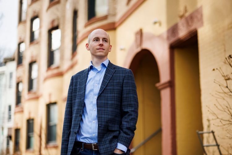 Portrait of Christopher Palmer PhD ’14 outside with city buildings behind him.