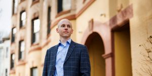 Portrait of Christopher Palmer PhD ’14 outside with city buildings behind him.