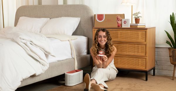 Loewen Cavill sits on the floor next to a bed holding a small device. There is a larger device on the floor next to her and a pad on the bed. Behind Cavill, a cardboard box rests on a chest of drawers.